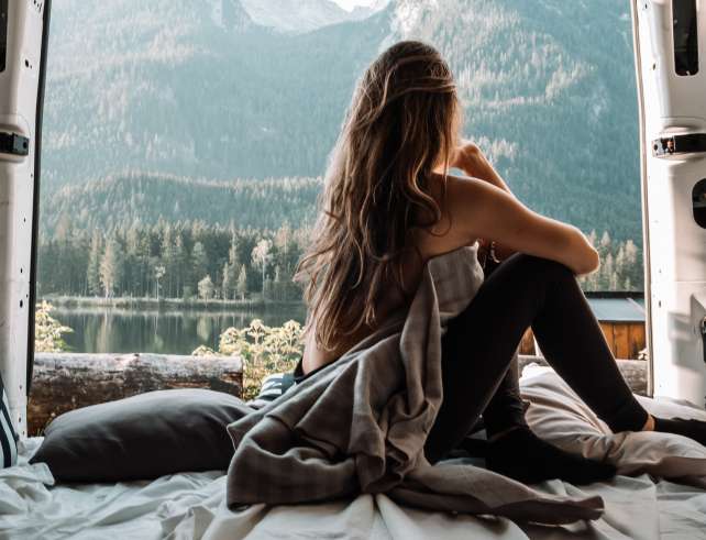 The picture displays a girl enjoying great views while just woken up from her bed inside a car to showcase coziness from out of a car